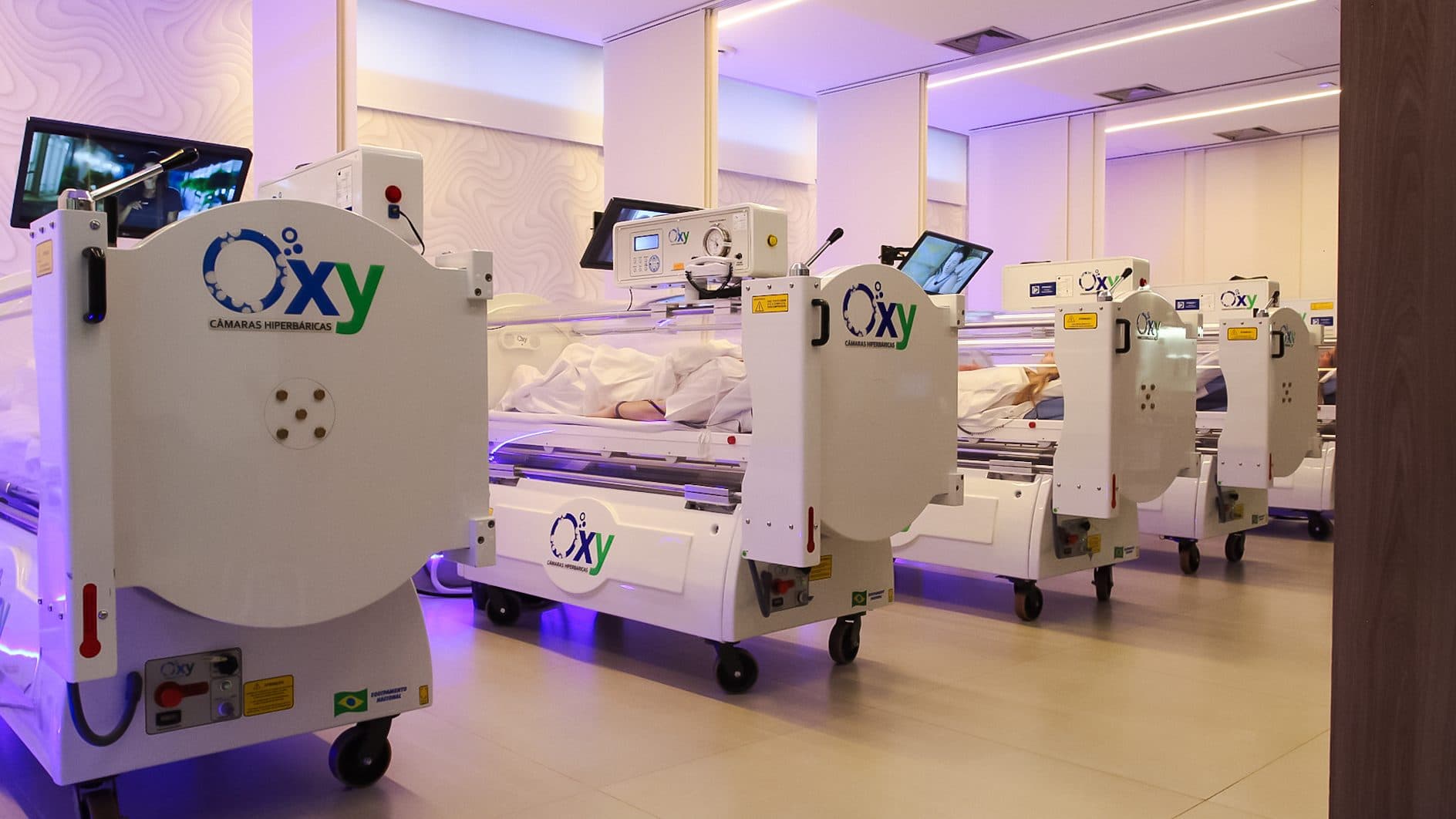 Fila de câmaras hiperbáricas Oxy alinhadas em sala com iluminação lilás, monitor superior em cada câmara e paciente sob lençol branco — ambiente clínico premium da Oxy Recovery.